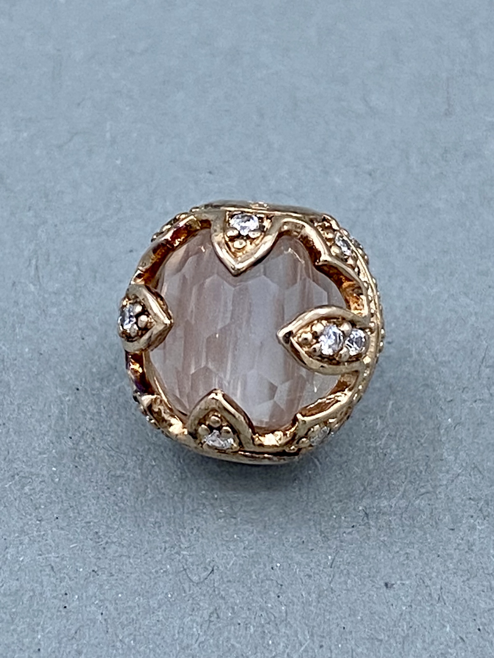 Ein kugelförmiger, goldener Anhänger mit einem rosa facettierten Stein und kleinen weißen Steinen, der von kleinen Blättern umrahmt wird.
