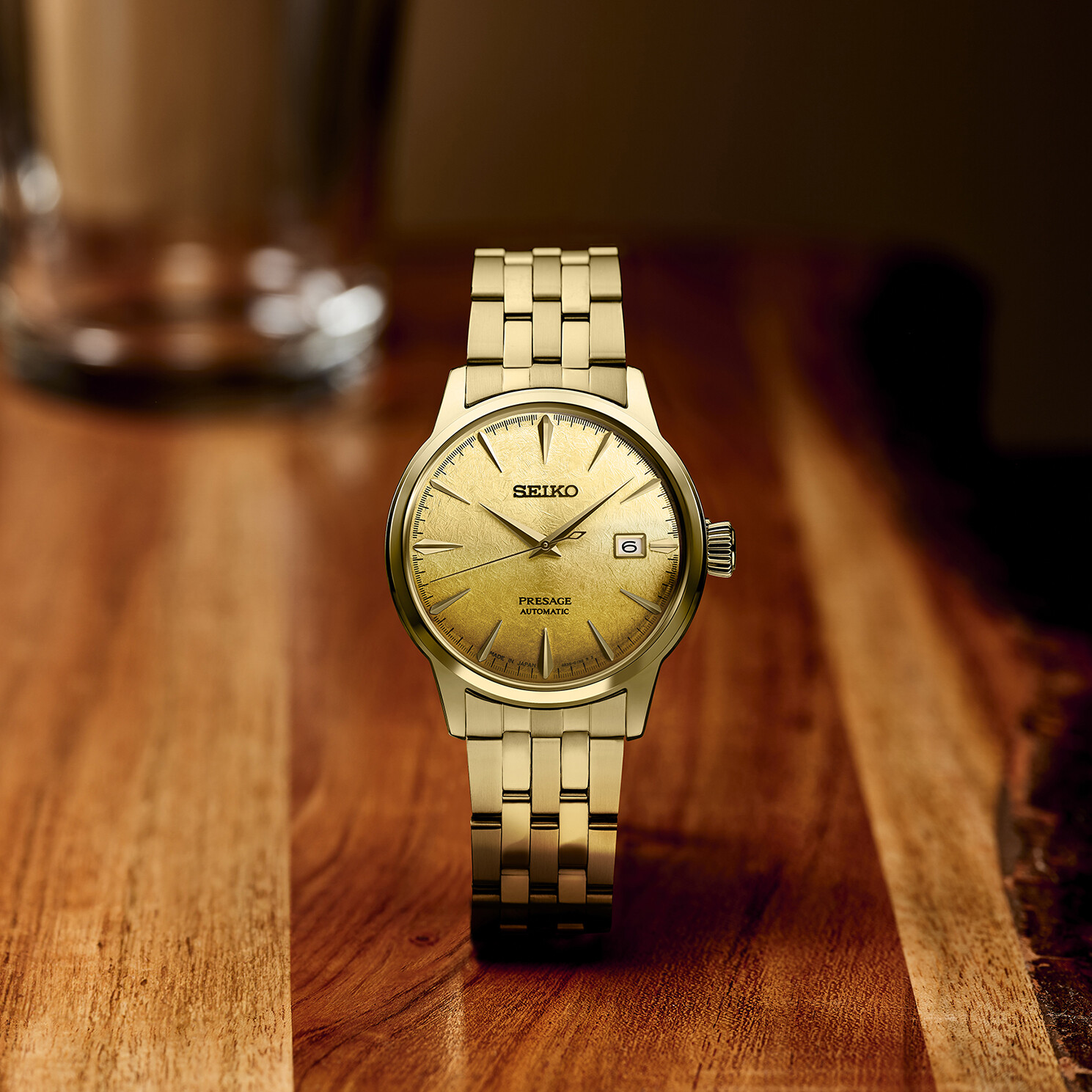 Ein goldfarbenes Armbanduhr mit einem runden Zifferblatt mit goldenem Muster und einem Datumsfenster liegt auf einem Holztisch.