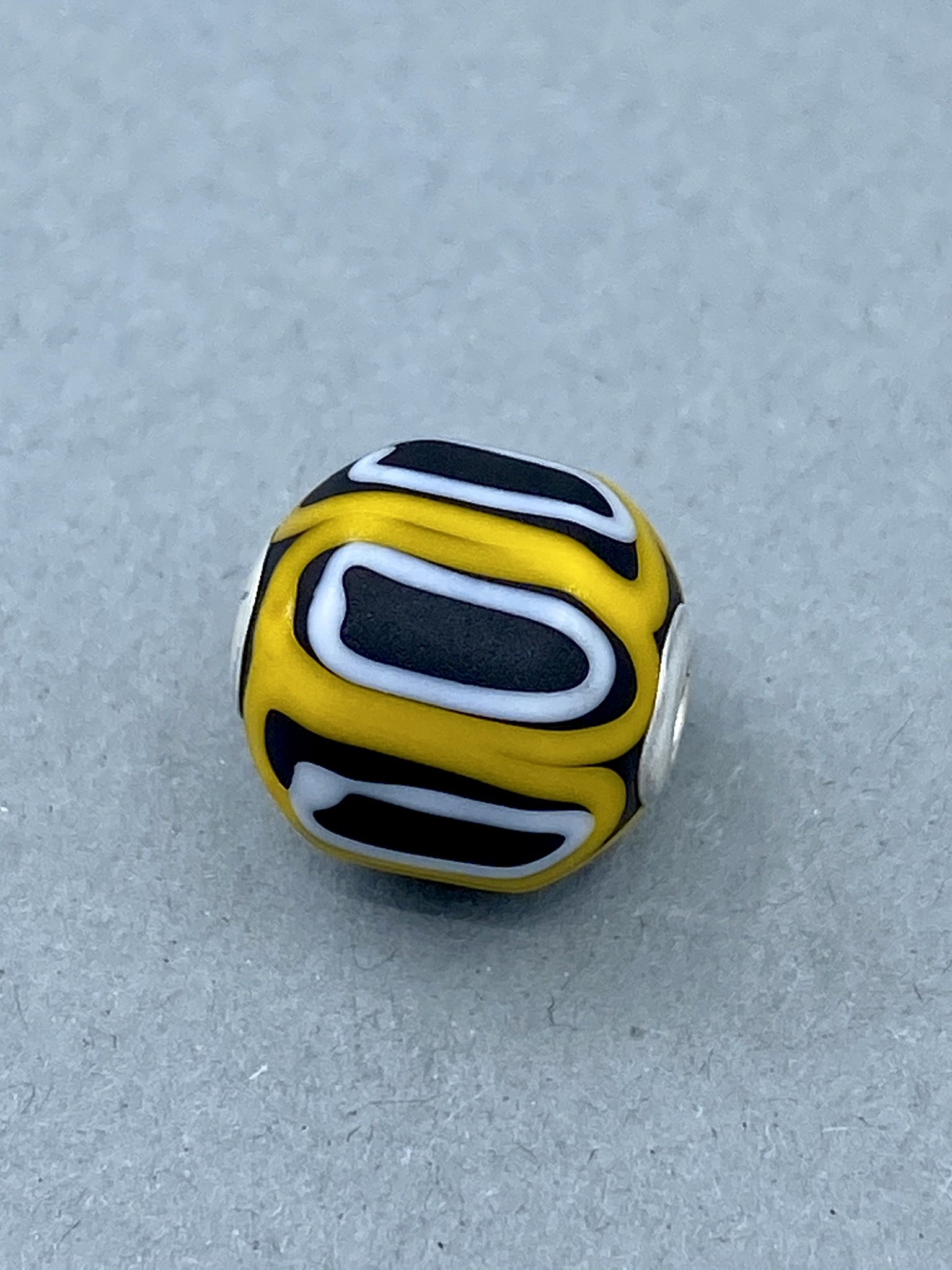 Eine runde, gelbe Perle mit schwarzen und weißen Linienmustern, die wie Wellen oder Augen aussehen.