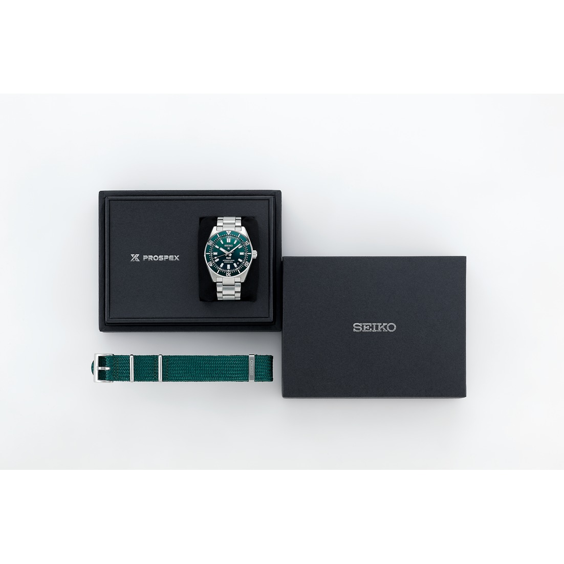 Grüne Uhr mit silbernem Armband in schwarzer Box neben schwarzer Geschenkbox und grünem Armband mit silbernen Streifen.