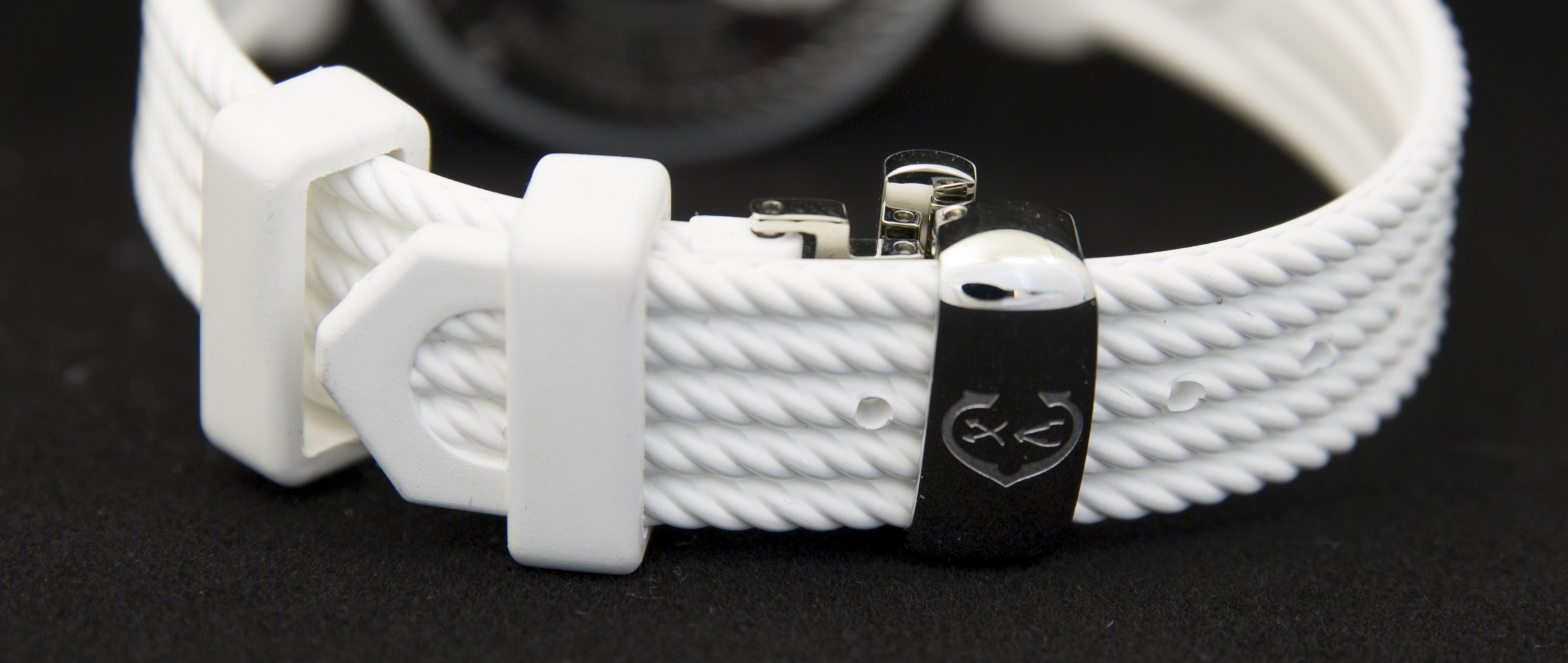 Ein weißes Armband mit geflochtenem Muster und einer schwarzen Schnalle mit einem Herzsymbol.