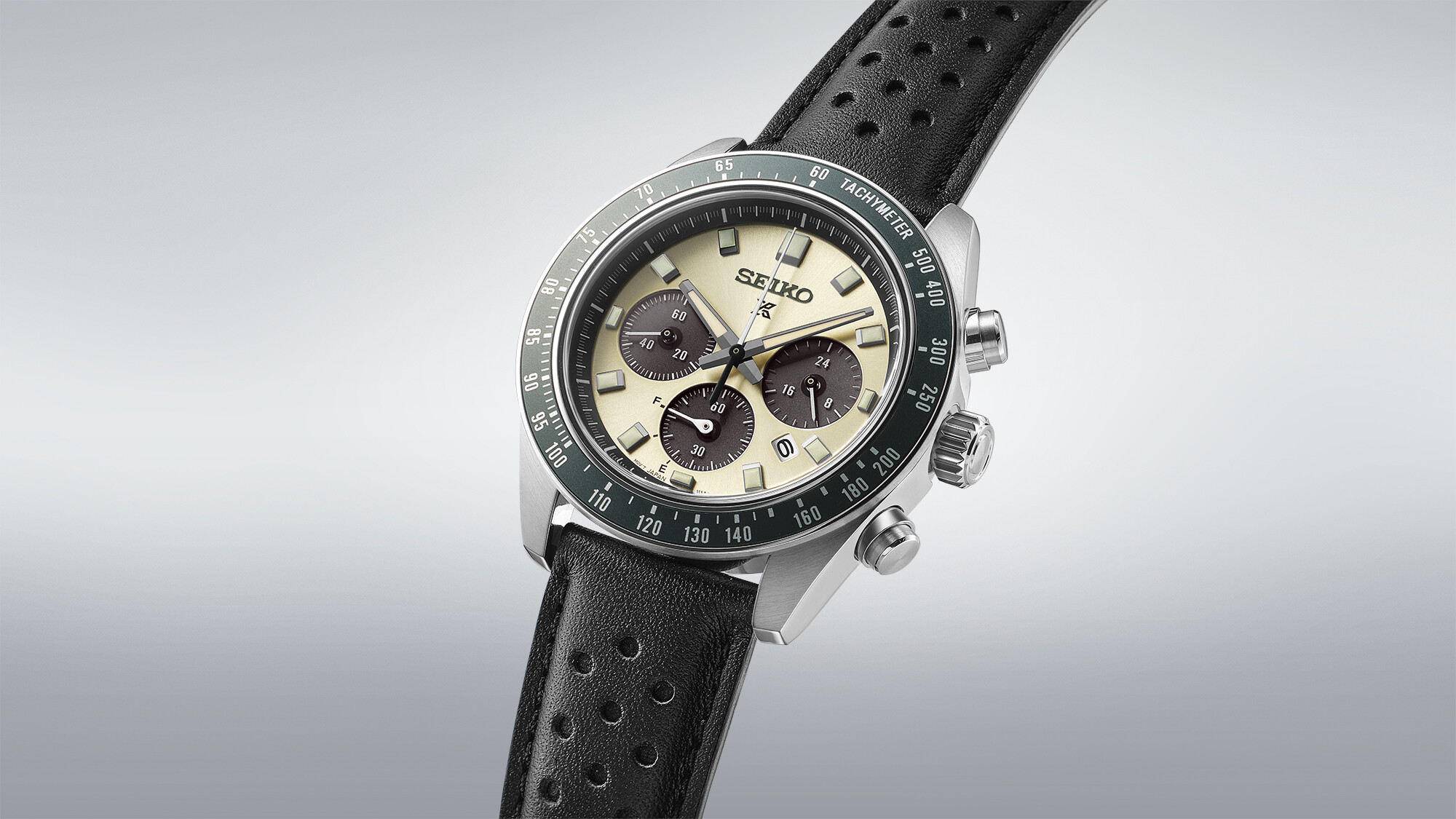 Ein Armbanduhr mit cremefarbenem Zifferblatt, schwarzen Zählern und grünem Tachymeter, befestigt an einem schwarzen Lederarmband.