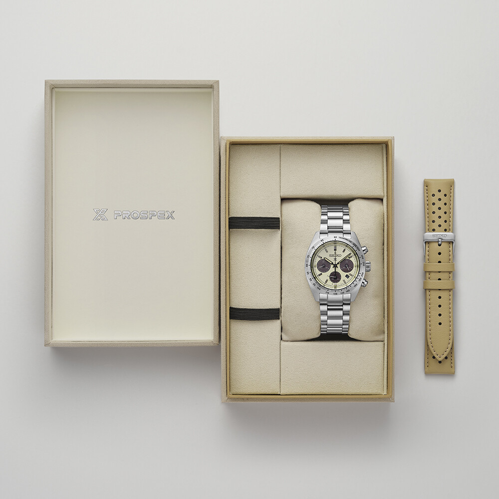 Ein silberner Chronograph mit cremefarbenem Zifferblatt und beigefarbenem Ersatzarmband liegt auf einem hellbeigen Hintergrund.