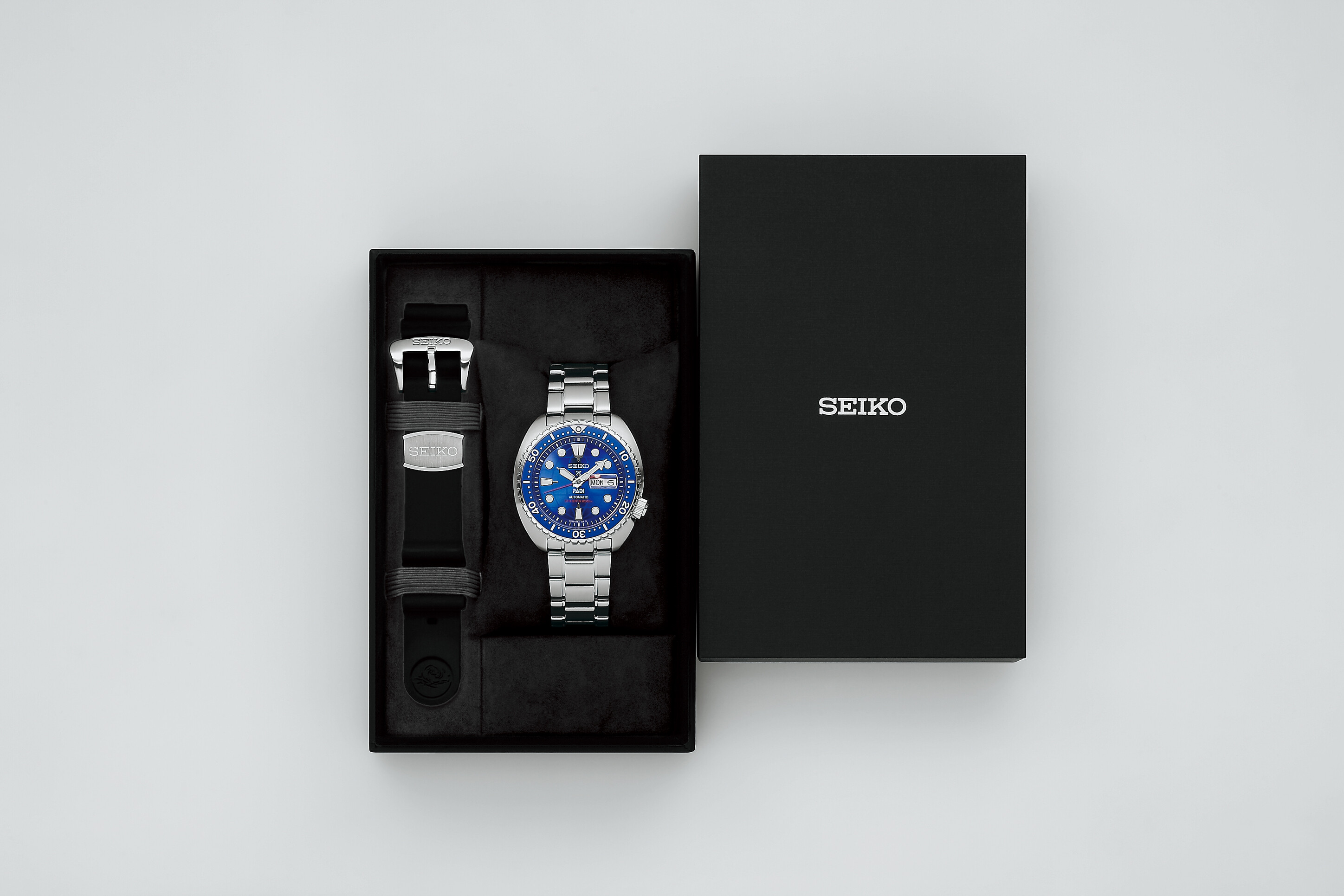 Eine silberne Herrenuhr mit blauem Zifferblatt liegt in einer schwarzen Box neben einem zusätzlichen schwarzen Armband.